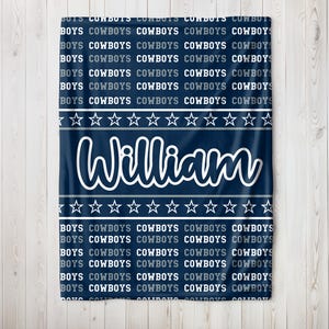 May include: A navy blue blanket with the name "William" in white script. The blanket features repeating text that says "BOYS COWBOYS" and a pattern of white stars. A personalized baby blanket.