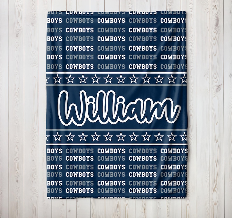 May include: A navy blue blanket with the name "William" in white script. The blanket features repeating text that says "BOYS COWBOYS" and a pattern of white stars. A personalized baby blanket.