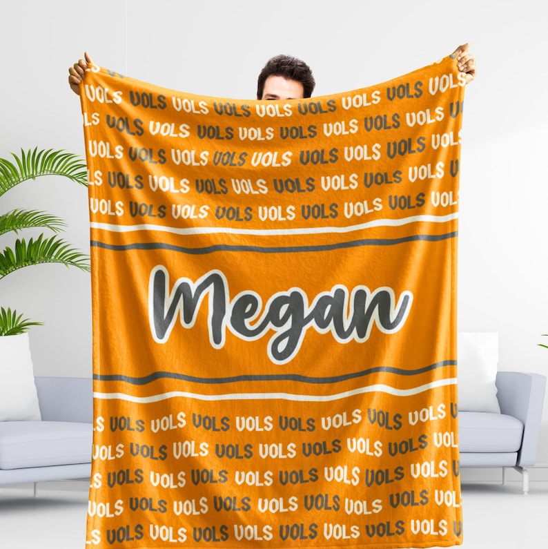 May include: An orange blanket with the word "Megan" in a large, stylized font. The blanket has a repeating pattern of the word "VOLS" in white and orange. Gray and white stripes are also present.