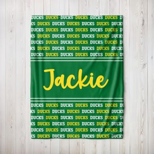 May include: Green blanket featuring the name "Jackie" in yellow script. The word "DUCKS" is printed repeatedly in yellow on a green background. White stripes border the name.