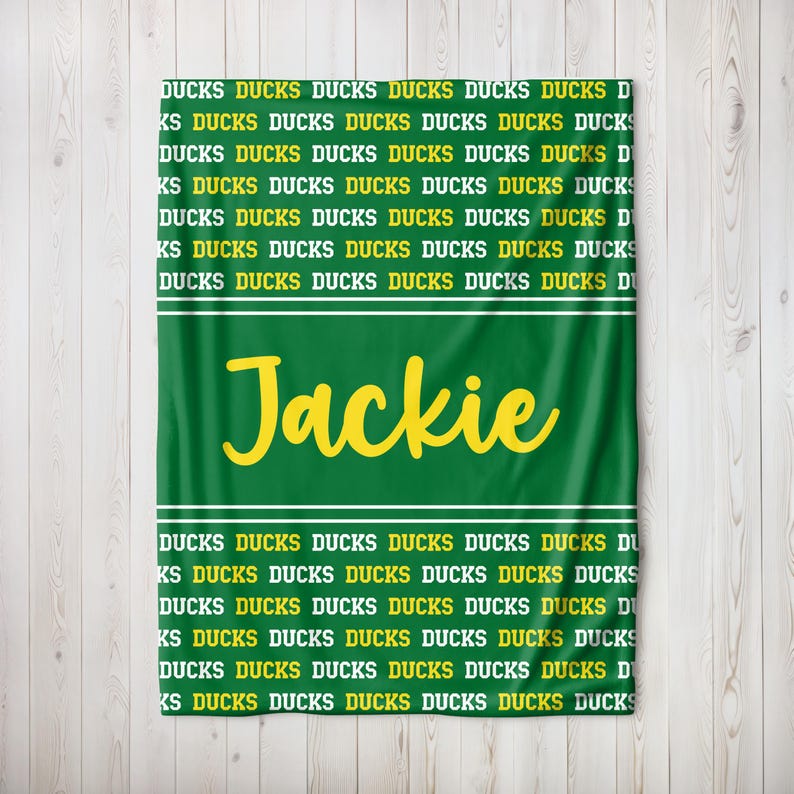 May include: Green blanket featuring the name "Jackie" in yellow script. The word "DUCKS" is printed repeatedly in yellow on a green background. White stripes border the name.