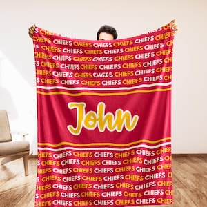 May include: Red and gold blanket featuring the word "Chiefs" repeated across the top and bottom. The name "John" is displayed in gold script in the center. A cozy, personalized blanket.