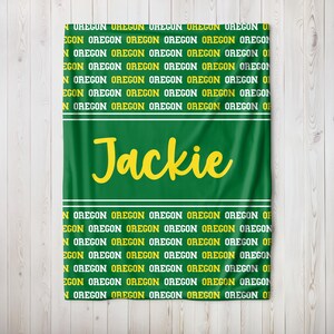 May include: Green blanket featuring the name "Jackie" in yellow script. The word "OREGON" is printed repeatedly in yellow and white, forming a pattern. White stripes border the name.