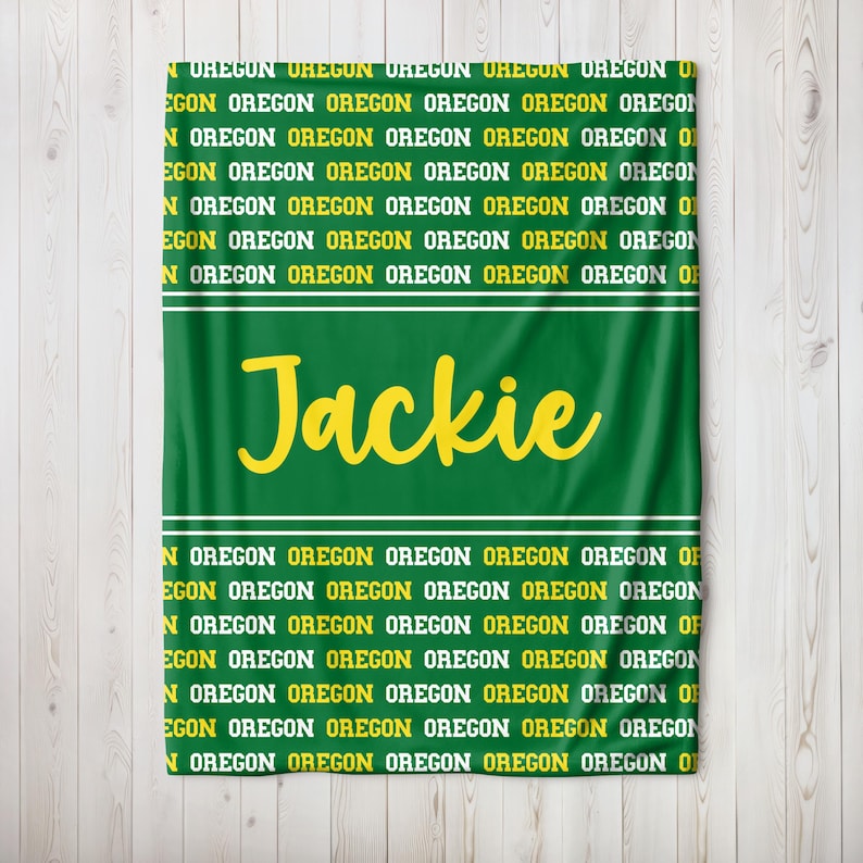 May include: Green blanket featuring the name "Jackie" in yellow script. The word "OREGON" is printed repeatedly in yellow and white, forming a pattern. White stripes border the name.
