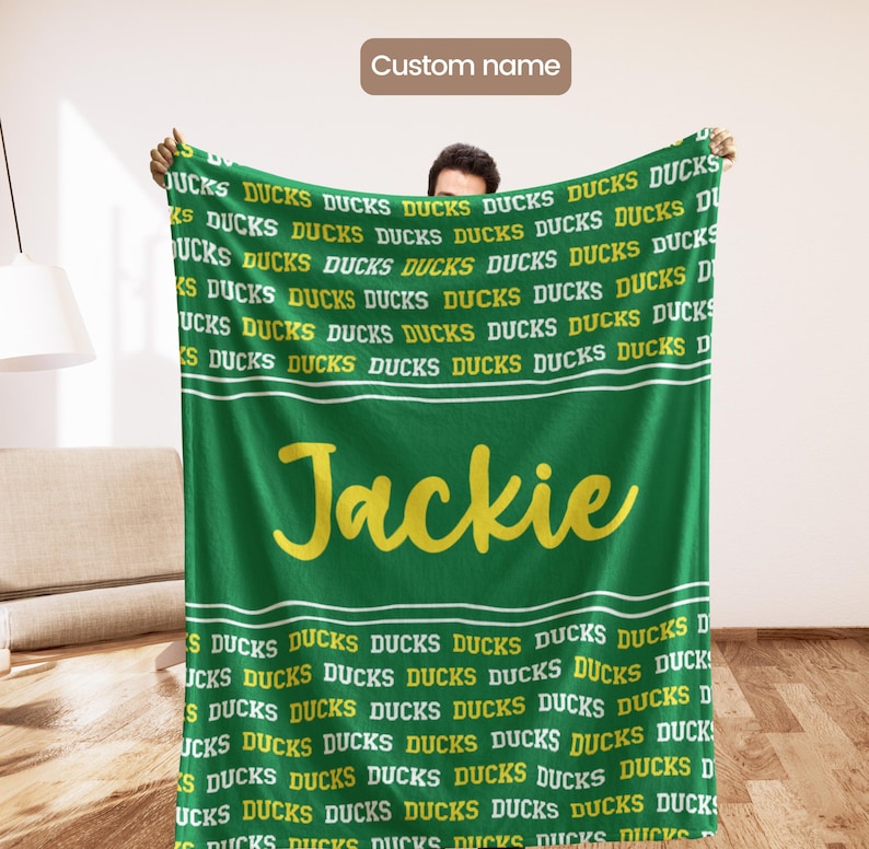 May include: A green blanket featuring the word "Ducks" in a repeating pattern, with the name "Jackie" in yellow script. The blanket has white stripes and is held up by a person.