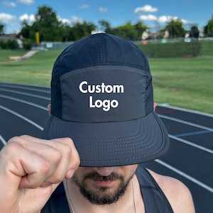 May include: A black five-panel cap with a flat brim. The cap features a rectangular patch on the front with the words "Custom Logo" in white. The cap is being held by a person in front of a track and field.
