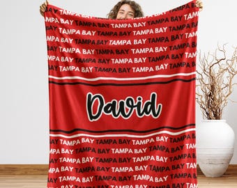 May include: Red blanket with the words "TAMPA BAY" in white repeating across the top and bottom. The name "David" is in the center in a script font. The blanket has black and white stripes.