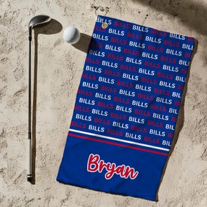 May include: A blue golf towel with the word "BILLS" in a repeating pattern. The bottom of the towel has the name "Bryan" in red script. A golf club and ball are next to the towel.
