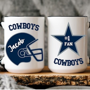 May include: Two white ceramic mugs with black handles and rims. One mug features a navy blue football helmet design with the name "Jacob" and the word "Cowboys". The other mug has a navy blue star with the text "#1 FAN" and "Cowboys".