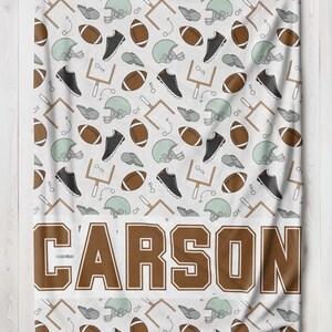 Op de afbeelding: Witte deken met een herhalend patroon van American football helmen, ballen, schoenplaatjes en speeldiagrammen. De naam "CARSON" staat in grote bruine letters onderaan.