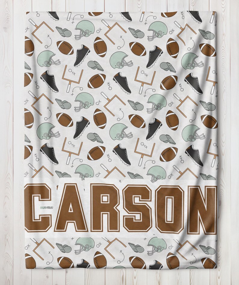 Op de afbeelding: Witte deken met een herhalend patroon van American football helmen, ballen, schoenplaatjes en speeldiagrammen. De naam "CARSON" staat in grote bruine letters onderaan.