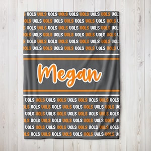 May include: A gray blanket with the word "Megan" in orange script. The blanket has an orange and white stripe design and is covered in the word "VOLS" in orange and white.