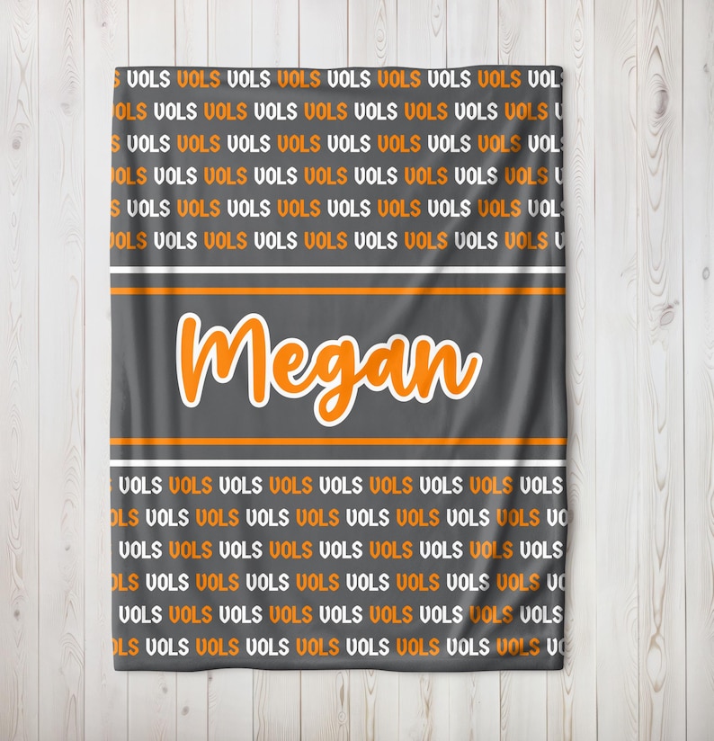 May include: A gray blanket with the word "Megan" in orange script. The blanket has an orange and white stripe design and is covered in the word "VOLS" in orange and white.