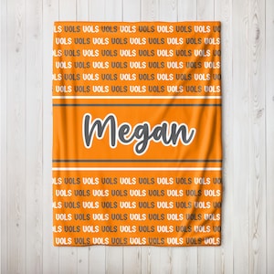 May include: Orange blanket with the name "Megan" in white and gray. The blanket has the word "VOLS" repeated throughout the design. Two gray and white stripes are above and below the name.
