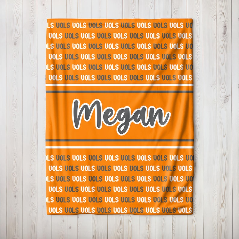 May include: Orange blanket with the name "Megan" in white and gray. The blanket has the word "VOLS" repeated throughout the design. Two gray and white stripes are above and below the name.