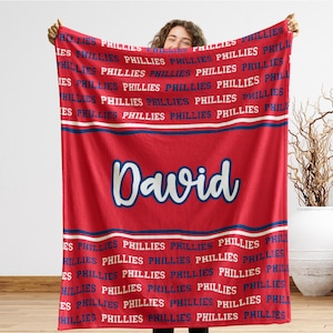 May include: A red blanket featuring the word "Phillies" in white and blue repeated across the top and bottom. The name "David" is displayed in a cursive font in the center. The blanket is held up by a person.