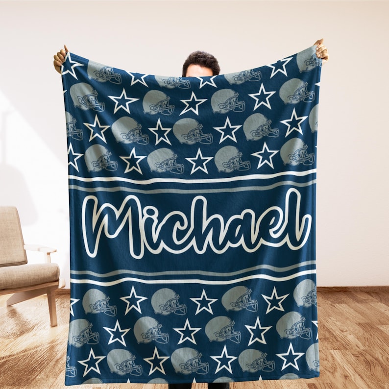 May include: A navy blue blanket featuring a pattern of football helmets and stars. The name "Michael" is written in white script across the center. The blanket is a cozy home decor item.