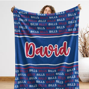 May include: A blue blanket with the word "BILLS" in red and white repeated across the top and bottom. The name "David" is displayed in a large, cursive font in the center. The blanket is held up by a person.