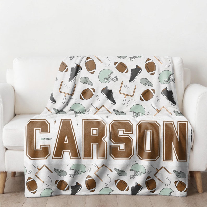Op de afbeelding: Een witte deken met een herhaald patroon van American footballs, helmen, schoenplaatjes en doelpalen. De naam "CARSON" staat in grote bruine letters onderaan de deken.
