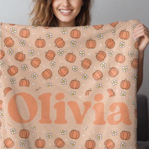 May include: A peach-colored blanket featuring a pattern of orange pumpkins and white flowers. The blanket has the name "Olivia" in large, orange, retro-style lettering. The blanket is soft and cozy, perfect for fall.