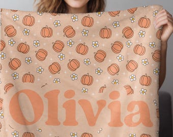 May include: A peach-colored blanket featuring a pattern of orange pumpkins and white flowers. The blanket has the name "Olivia" in large, orange, retro-style lettering. The blanket is soft and cozy, perfect for fall.