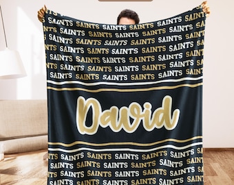 May include: Black blanket with the word "Saints" in gold and white repeated across the top and bottom. The name "David" is displayed in large gold letters in the center. The blanket is held up by a person.