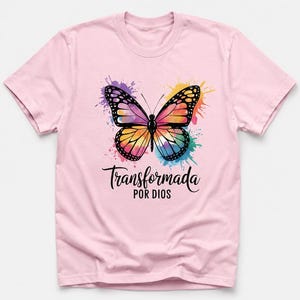 May include: A light pink t-shirt featuring a central graphic of a butterfly with multicolored watercolor-style wings. Below the butterfly, the black text reads "Transformada POR DIOS" in a combination of script and sans-serif fonts.