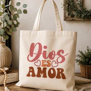 Op de afbeelding: Crèmekleurige draagtas met de tekst "Dios es Amor" in roze en bruine tinten. De tas heeft twee handvatten en is gemaakt van stevig canvas materiaal. Het ontwerp bevat hartvormen.