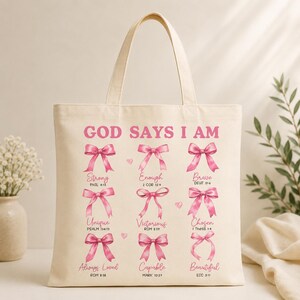 Puede incluir: Bolso tote color crema con la frase "GOD SAYS I AM" en rosa. Debajo, la bolsa presenta lazos rosas con palabras como "Fuerte", "Suficiente" y "Valiente". Diseño sencillo con asa.