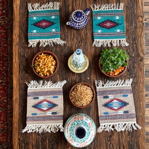 May include: A rustic wooden table displays a collection of decorative items. Six woven placemats with fringed edges and geometric patterns in teal, beige, and red are arranged around small bowls of food and a blue teapot.