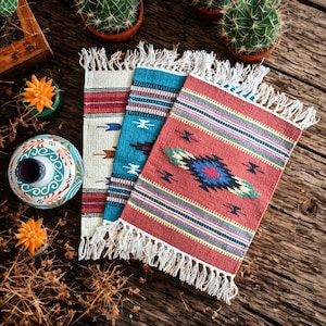 May include: Three small, rectangular woven rugs with fringed edges. The rugs feature colourful geometric patterns in red, turquoise, and cream. The rugs are displayed on a rustic wooden surface, alongside decorative items and potted cacti.