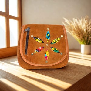 Può includere: Borsa a tracolla in camoscio marrone con un design floreale. La borsa ha una chiusura con cerniera e borchie decorative. Il design floreale è realizzato in tessuto ricamato colorato. La borsa è su una superficie di legno.
