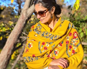 Puede incluir: Un chal de color amarillo mostaza con intrincados bordados florales en tonos rojo, azul y blanco. La prenda presenta patrones de costura detallados y un borde con flecos, lo que le otorga una apariencia texturizada a la superficie.