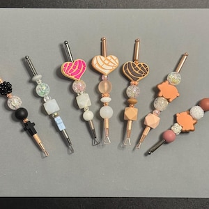 May include: A collection of seven decorative tools with copper-colored metal shafts. Each tool features a unique design of beads, geometric shapes, and heart or pig-shaped accents in various colors, including pink, white, and black. The tools are arranged on a gray surface.