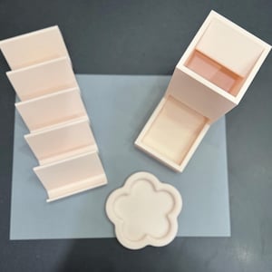 May include: A light peach-colored desk organizer set consisting of a tiered card holder with five slots, a tall rectangular pen holder with an attached base tray, and a small flower-shaped coaster, all arranged on a flat rectangular surface.