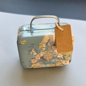 May include: A small, blue metal suitcase with a map of Europe printed on its sides. The suitcase has a silver handle and a blank, brown tag attached. The map includes countries like Iceland, Norway, and France.