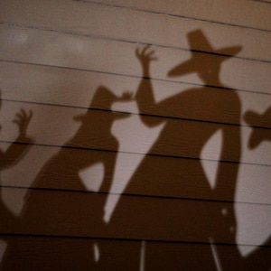 May include: Dark brown silhouettes of four figures wearing wide-brimmed hats cast onto a light brown horizontal paneled surface. The figures appear to be in motion with raised arms and distinct hat shapes, creating a dramatic shadow play effect.