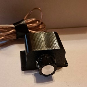 May include: A black electronic component with a knob and attached wires. The component has a rectangular body with a rounded knob on the front. The wires are copper-colored and wrapped together with black tape.