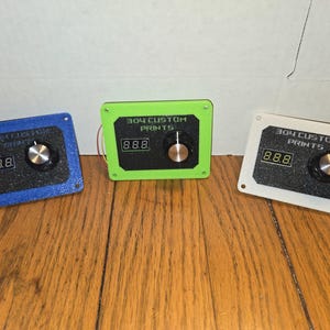 May include: Three custom-printed electronic devices in blue, green, and white. Each device has a digital display showing "8.8.8", a knob, and the text "304 CUSTOM PRINTS". The devices are on a wooden surface.
