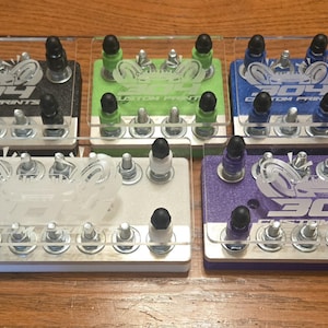 May include: A collection of colorful display stands with the text "304 Custom Prints". Each stand features a different color: black, green, blue, red, white, and purple. The stands have clear acrylic tops and are on a wooden surface.