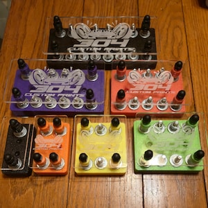 May include: A collection of custom printed display stands in various colors, including black, purple, orange, yellow, and green. Each stand features a clear acrylic top with the text "304 CUSTOM PRINTS" and multiple posts for holding items.