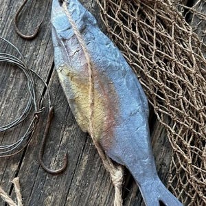 May include: A gray, textured fish-shaped ornament with a gold and copper-toned midsection, suspended by a piece of twine. The fish is accompanied by a rusty metal fishing hook with a wooden float and a length of coiled wire fishing line.