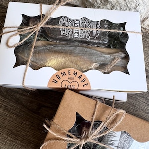 May include: A white rectangular gift box with a scalloped window containing a dark bottle labeled WHISKY and a gold-toned fish-shaped item, tied with twine and a sticker reading HOMEMADE WITH LOVE, alongside a partial brown cardboard box.