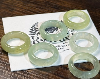 May include: Six translucent jade rings in varying shades of green are arranged on a white card with a fern logo and text. The rings are smooth and polished, showcasing the natural variations in the stone. The rings are a light green color.