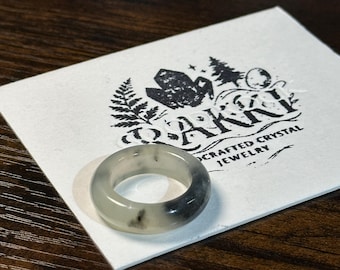 May include: A translucent ring with gray and black marbling sits on a white card with a logo that reads "DARKKI Crafted Crystal Jewelry". The ring is made of a smooth, polished material and is set against a dark wood grain surface.