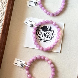May include: Three Kunzite crystal bead bracelets in varying sizes. The bracelets are a soft lavender color and are labeled 8mm, 9mm, and 10mm. The bracelets are displayed on a light beige surface, with a tag from the brand RAKKI.