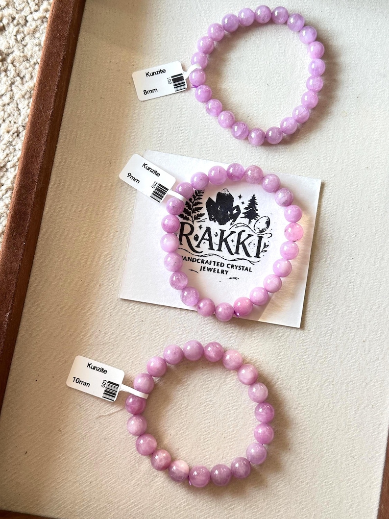 May include: Three Kunzite crystal bead bracelets in varying sizes. The bracelets are a soft lavender color and are labeled 8mm, 9mm, and 10mm. The bracelets are displayed on a light beige surface, with a tag from the brand RAKKI.