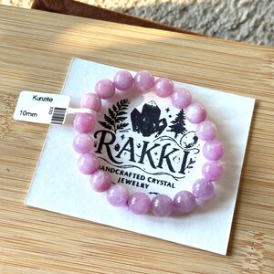 May include: A light purple Kunzite bracelet with 10mm beads. The bracelet is displayed on a white card with the brand name "Rakki" and the words "Handcrafted Crystal Jewelry". The card is on a wooden surface.