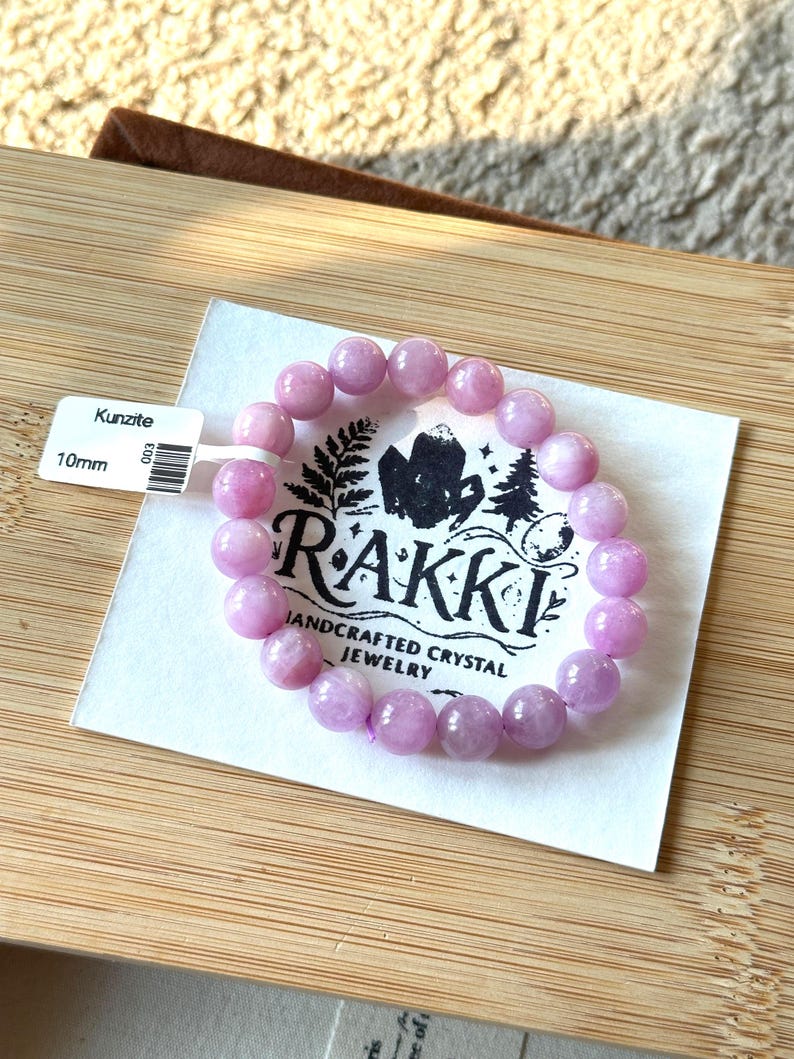 May include: A light purple Kunzite bracelet with 10mm beads. The bracelet is displayed on a white card with the brand name "Rakki" and the words "Handcrafted Crystal Jewelry". The card is on a wooden surface.