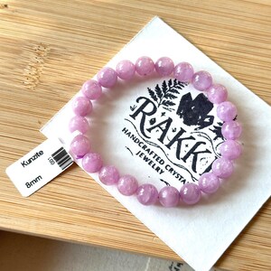 May include: A light purple kunzite bracelet with 8mm beads rests on a white paper with the brand name "Rakk" and the words "Handcrafted Crystal Jewelry". The bracelet is displayed on a wooden surface.